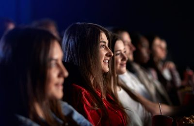 Side view of young stunning brunette girl watching movie in cinema, wearing casual outfit. Selective focus of caucasian girl smiling with teeth enjoying film with friends. Entertainment concept.